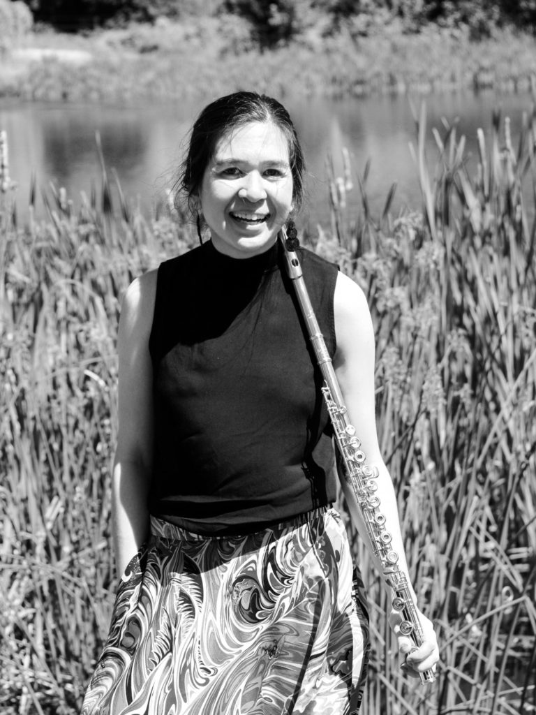 Black and white photo of Nicole, a Filipino-American flutist, standing at the shore of a small lake, in front of a cluster of tall grasses.  She smiles at the camera, holding her flute vertically in her left hand and resting it against her shoulder.  She wears a black sleeveless blouse and a white skirt with a swirling wave pattern, and her hair is pulled back in a low braided bun.  