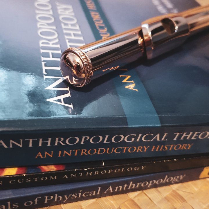 A rose gold flute headjoint lays diagonally on top of a stack of blue anthropology textbooks.  The titles on their spines read "Anthropological Theory: An Introductory History," "Custom Anthropology," and "Essentials of Physical Anthropology." 