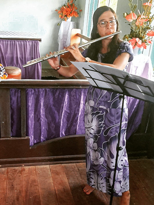 Nicole, a Filipino-American flutist, playing her flute in a church in Fiji.  Purple curtains hang from low railings behind her, and clusters of orange flowers border the window on her left.  She wears a purple sulu jaba with a pattern of white hibiscus flowers.  A black wire music stand is in front of her, at the right side of the photo.  