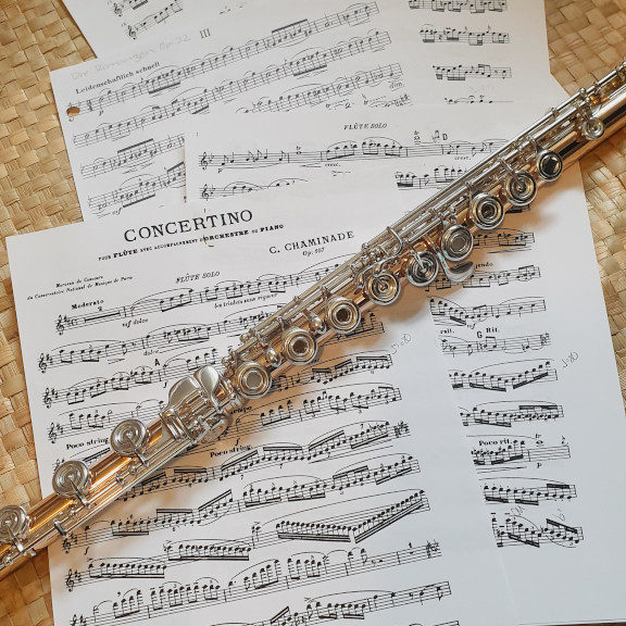 Sheets of flute music by Cecile Chaminade and Clara Schumann are spread out across a woven pandanus leaf mat. A gold flute with silver keys lies diagonally across the pages.