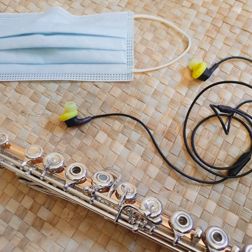 A blue face mask, yellow earbuds with a black cord, and a metal transverse flute lay across a pandanus leaf mat.  