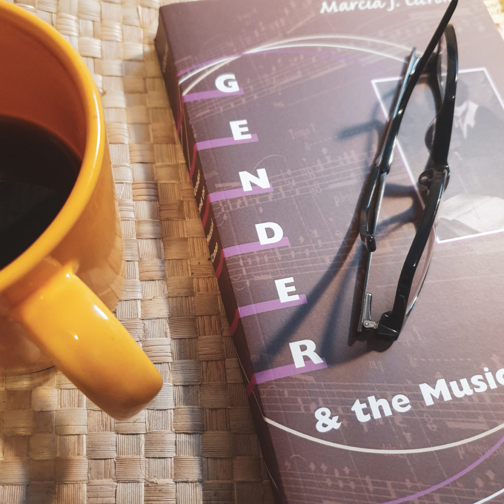 A copy of Marcia Citron's book "Gender and the Musical Canon," with a folded pair of black-framed eyeglasses sitting on top. A yellow mug of coffee sits next to it, half outside the photo's frame.